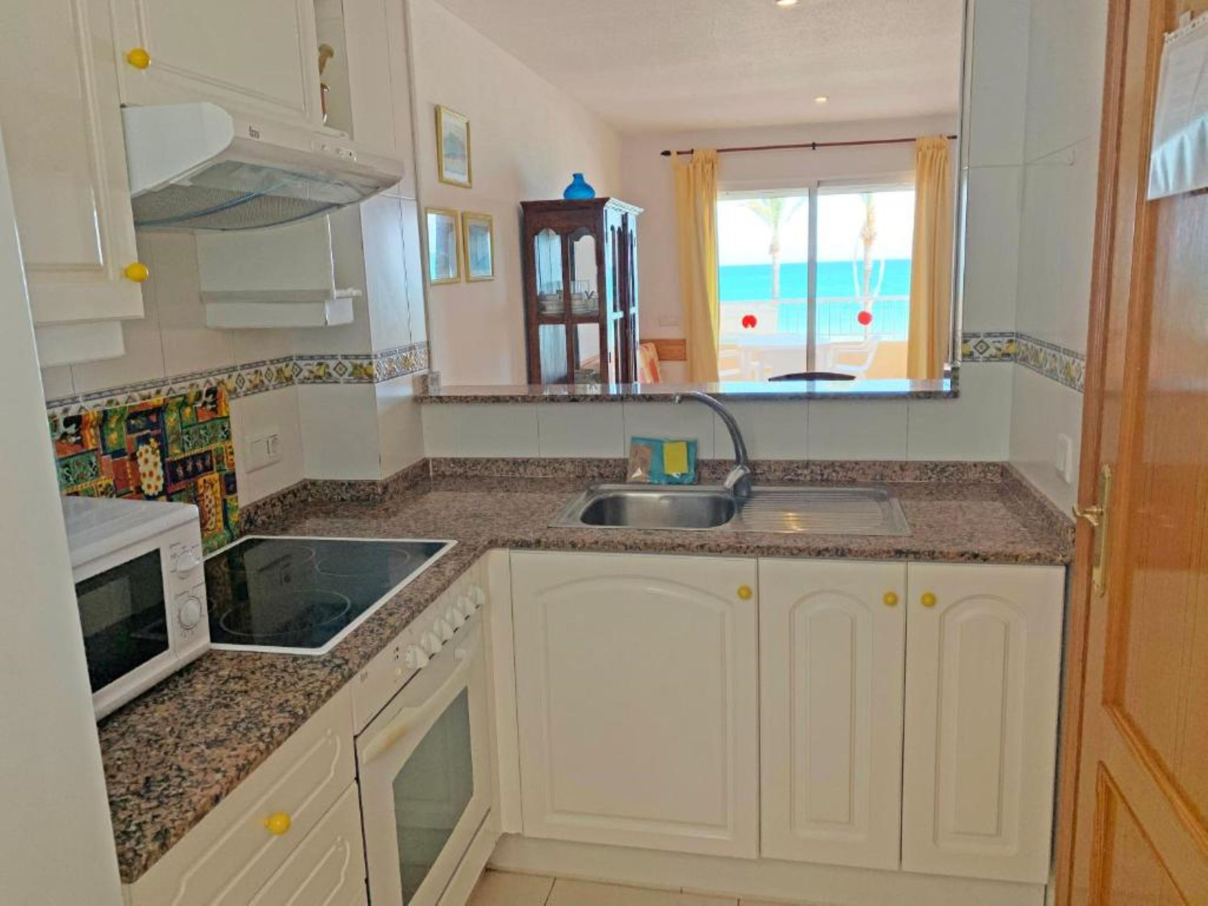 A compact kitchen with white cabinets, a granite countertop, a built-in oven, and a window with a view of the beach.