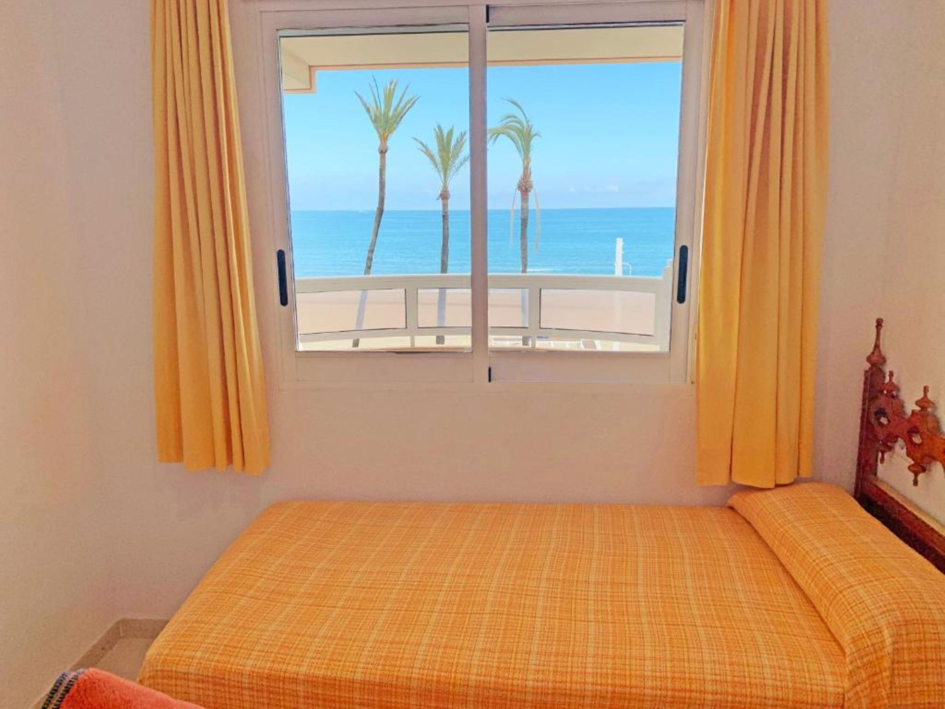 A room with a bed, a window with curtains, and a view of the sea and palm trees.