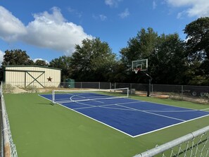 Una cancha de baloncesto al aire libre con superficie azul, valla metálica y árboles de fondo.