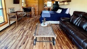 Ein gemütliches Blockhaus-Interieur mit Holzbalkendecke, einem Bett mit blauer Bettwäsche, einem Ledersofa und einem kleinen Esstisch.