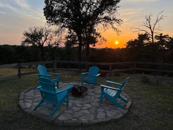 Un patio con cuatro sillas Adirondack azules dispuestas alrededor de un brasero, con el bosque y una puesta de sol como fondo.