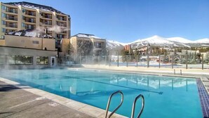 A swimming pool with clear blue water, surrounded by a building with multiple balconies and a view of snow-capped mountains in the background. - 3 Bedroom Townhome in Downtown Breckenridge, off Main Street With Pool, and Hot Tubs (Breckenridge)