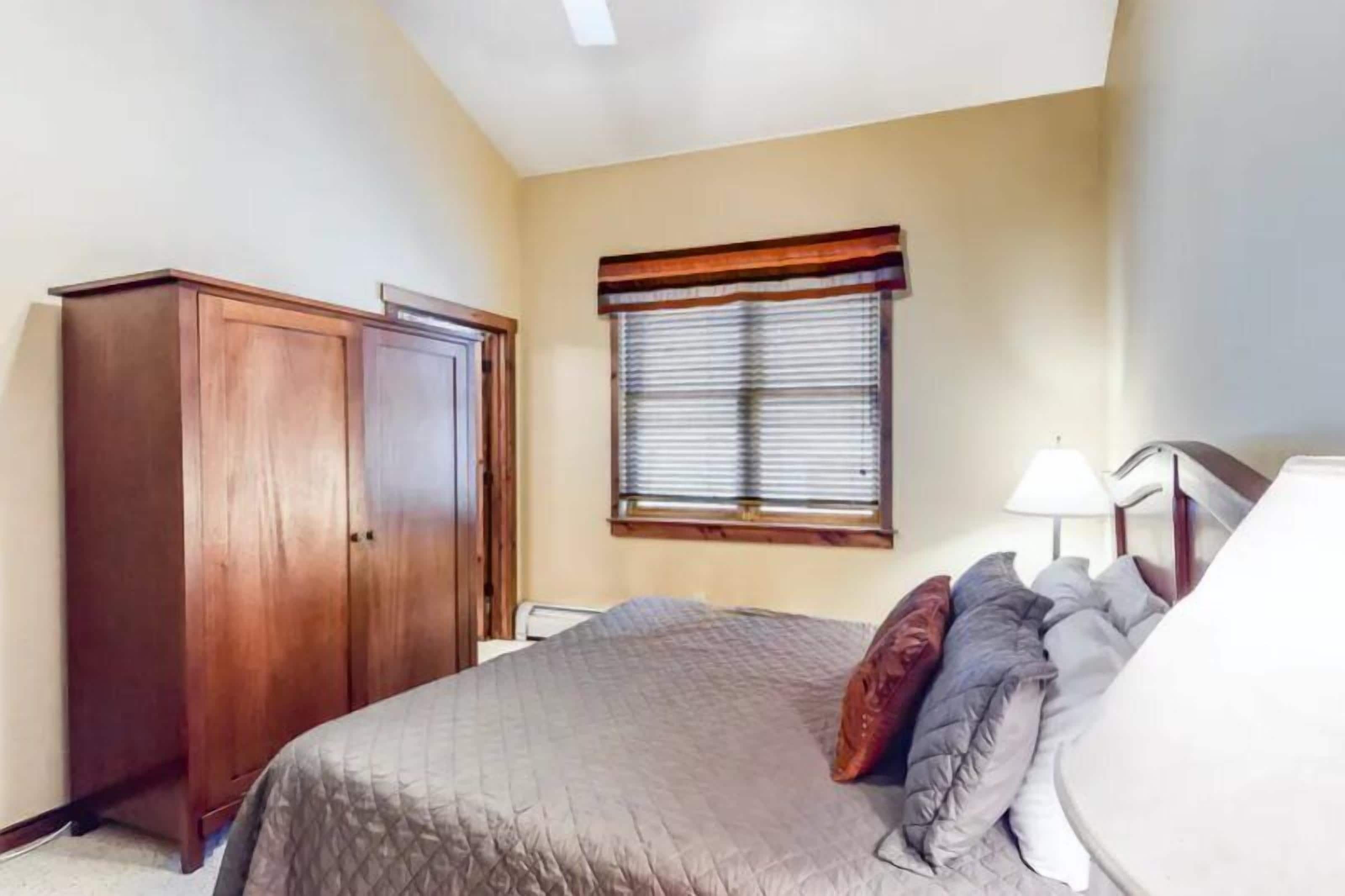 A bedroom with a bed, a wardrobe, a window with blinds, and a lamp.