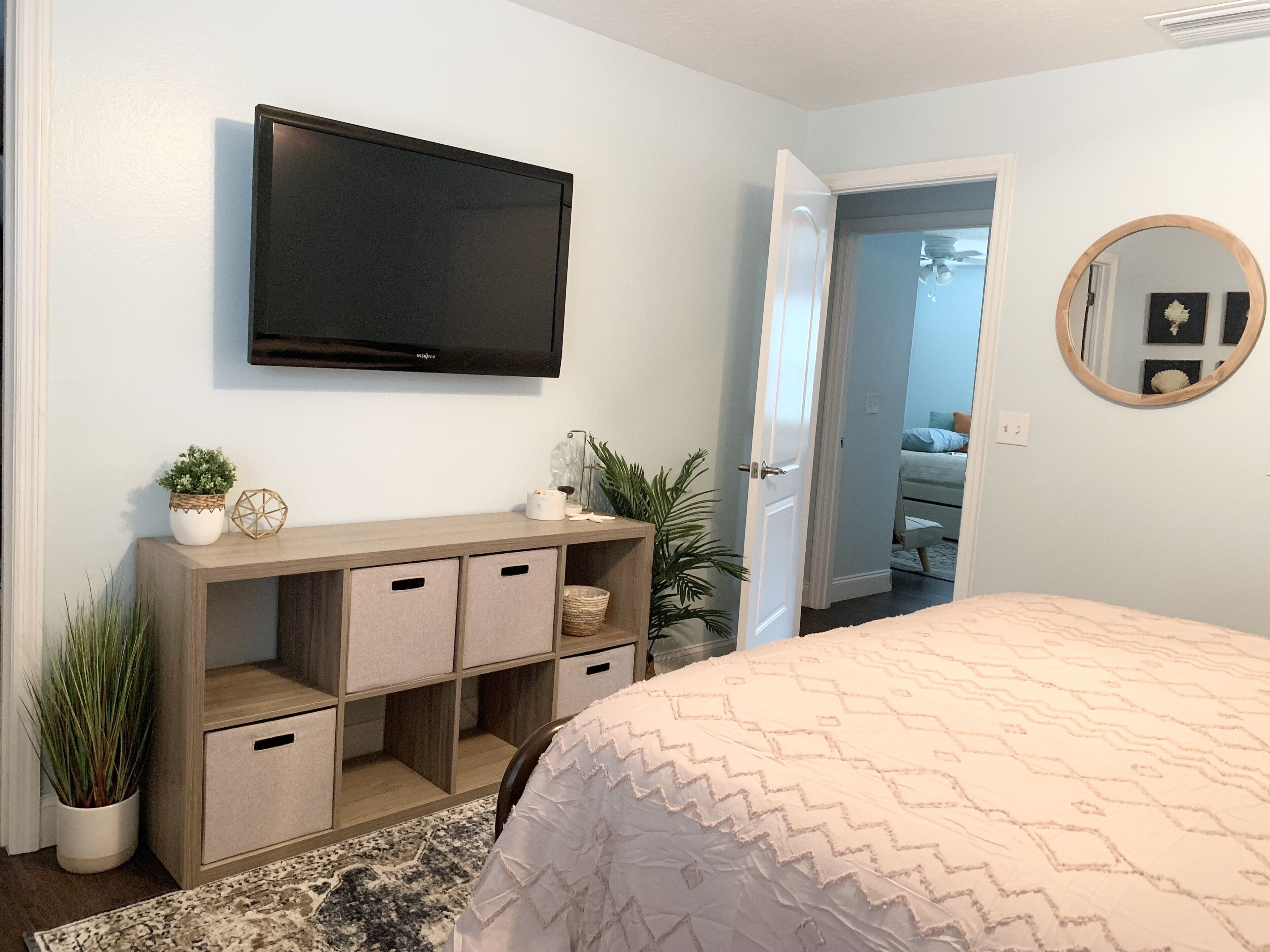 Une chambre à coucher comprenant un lit, un téléviseur fixé au mur, une console en bois avec des compartiments de rangement, un miroir et une porte donnant sur une autre pièce.