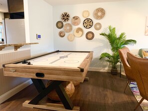 A pool table in a room with a wooden floor, a wall with woven art pieces, a potted plant, and a chair.