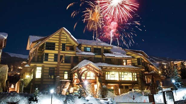 A large lodge with multiple levels, snow-covered surroundings, and a view of fireworks in the background.