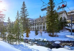 A ski resort with a building surrounded by snow-covered trees and a stream. - 2 Bedroom Platinum Condo Near Chairlift in Cascade Village With Pool and Hot Tub (Vail)