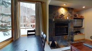 A living room with a stone fireplace, a dining table, and a view of a snowy outdoor area. - 2 Bedroom Platinum Condo Near Chairlift in Cascade Village With Pool and Hot Tub (Vail)