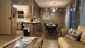 A cozy living room with a leather sofa, a coffee table with magazines, a dining area with a table and chairs, and a kitchen with wooden cabinets and a window.