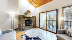 A modern living room with a stone fireplace, a round coffee table, and a large sliding glass door leading to a balcony. - Chic Ski In, Ski Out 2 Bedroom Penthouse in Beaver Creek Village (Avon)
