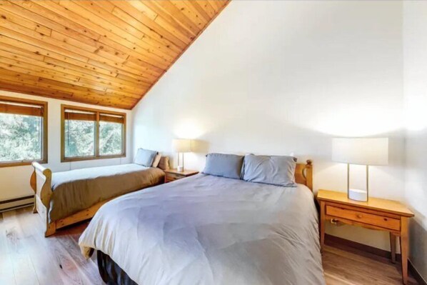 A bedroom with a sloped ceiling, two beds, a nightstand with a lamp, and large windows.