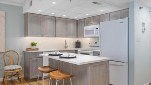 A modern kitchen with grey cabinets, a white island with stools, a built-in microwave, and a refrigerator. - LAST DATES! Bay Sunset Getaway • Pool • 2BR/2BA • Free Linens (Ocean City)