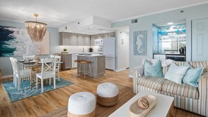A modern living room with a striped sofa, a dining table with chairs, and a kitchen area with a central island. - LAST DATES! Bay Sunset Getaway • Pool • 2BR/2BA • Free Linens (Ocean City)