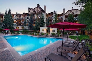 A swimming pool with lounge chairs and umbrellas, surrounded by multi-story buildings with balconies and trees.