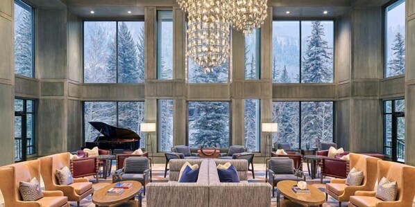 Enjoy expansive views of the snowy forest from the large windows in this elegantly furnished living area, featuring a grand piano and comfortable seating.