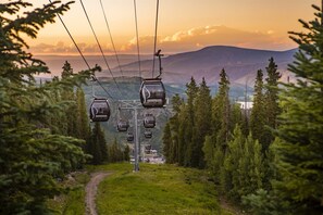 A ski lift with cabins labeled "Snowmass" against a backdrop of mountains and trees during sunset. - Luxury Ski in, Ski out 1 Bedroom Colorado Resort Vacation Rental in the Heart of Snowmass Base Village (Snowmass Village)