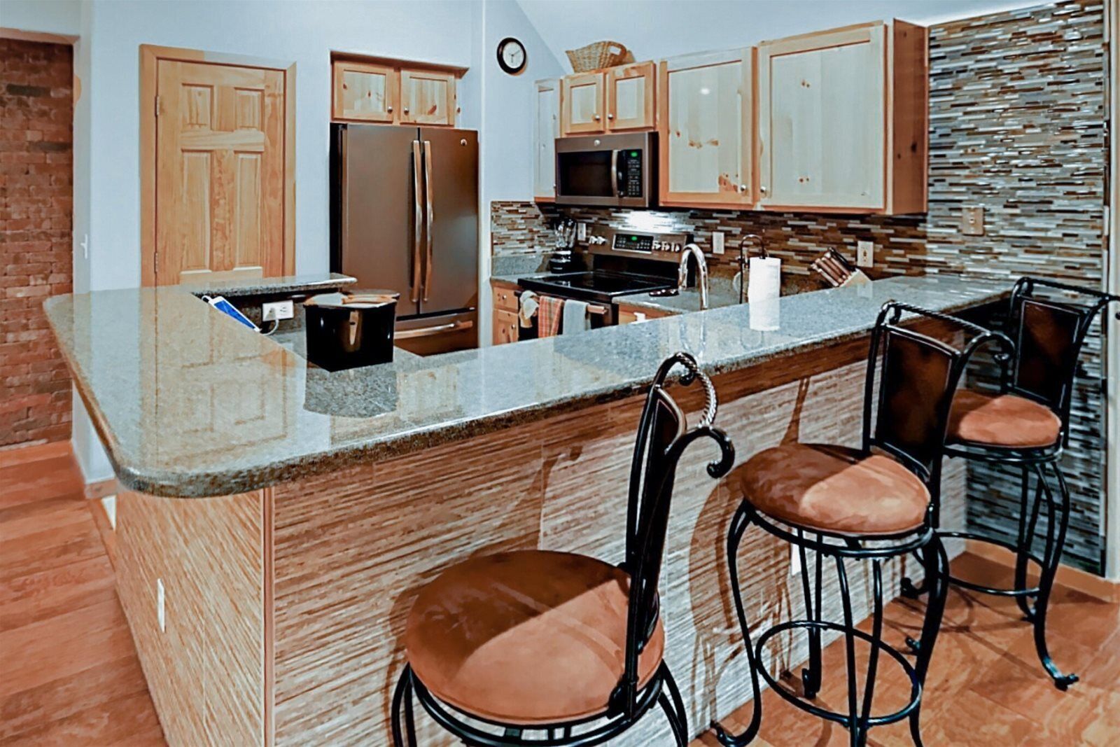 A kitchen with wooden cabinets, a stainless steel refrigerator, a microwave, and a stone backsplash. There are three bar stools with cushioned seats.