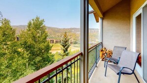 A balcony with two chairs, a stack of firewood, and a view of trees and buildings in the distance. - Premier Ski in, Ski-out 3 Bedroom Condo With Spectacular Views, Heated Outdoor Pool and Hot Tub, Short Walk to Village (Snowmass Village)