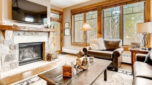 A cozy living room with a fireplace, a large TV, comfortable seating, and large windows. - Luxury 2 Bedroom Mountain Vacation Residence With Hot Tub, Pool, and Easy Access to Ski Slopes (Breckenridge)