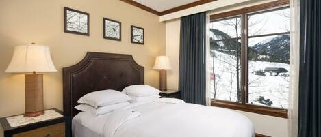 Una habitación de hotel con cama, dos lámparas de noche, una mesita de noche con cajones, una ventana con cortinas y tres cuadros en la pared.