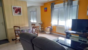 A living room with a sofa, a TV, a dining table with chairs, and a window with blinds. - La douceur de vivre (Blois)