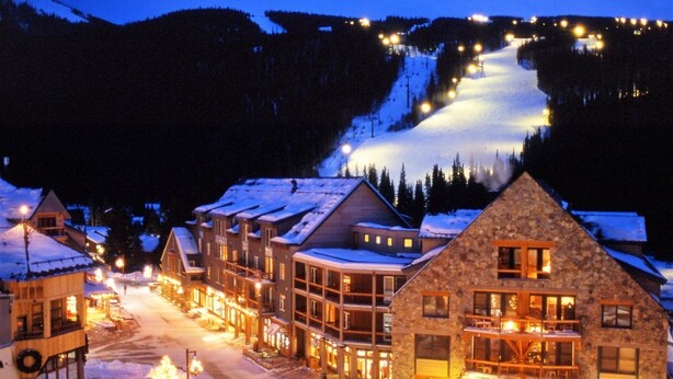 A snow-covered village with wooden buildings, a ski slope in the background, and a lit street.