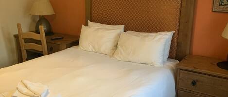 Una cama bien hecha con sábanas y almohadas blancas, un cabecero de madera, una mesita de noche con lámpara y un tocador con reloj.