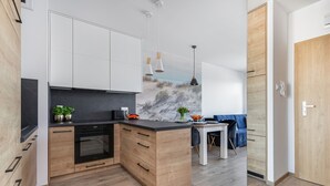 A modern kitchen with wooden cabinets, a black countertop, and a built-in oven. There is a dining area with a table and chairs, and a blue sofa. - Seaside Apartamenty Kasprowicza (Kolobrzeg)