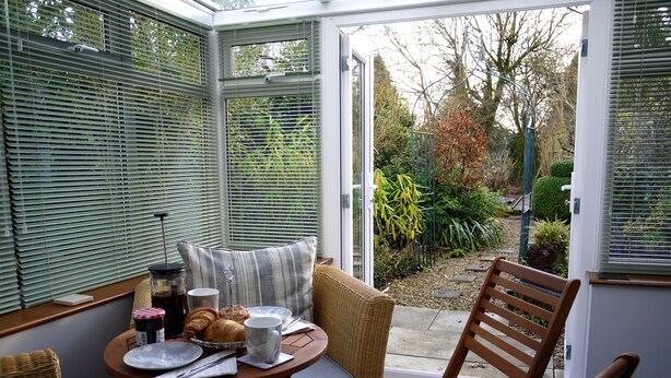 A conservatory with a dining table set for two, surrounded by wicker furniture, and a view of the garden outside. - Cosy Stone Cottage - The Bungalow Perrotts Brook - Detached, parking, wifi. (Perrotts Brook Cirencester)