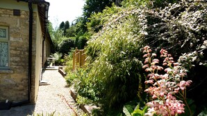 A garden pathway leading to a stone building with a window displaying a plant. - Cosy Stone Cottage - The Bungalow Perrotts Brook - Detached, parking, wifi. (Perrotts Brook Cirencester)