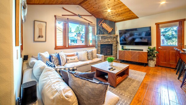 Un salon chaleureux avec un plafond en bois, une cheminée en pierre, une grande télévision et un canapé agrémenté de nombreux coussins.
