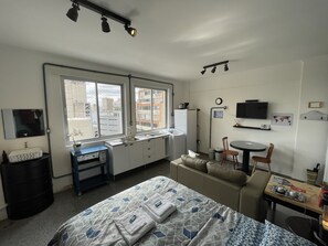 A compact hotel room with a bed, a small desk, a chair, a TV, and a window with a view of buildings.
