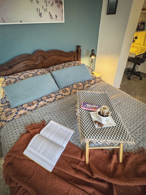 Une chambre à coucher avec un lit, une table de chevet sur laquelle se trouvent un livre et une collation, une lampe et un mur orné d’un tableau.
