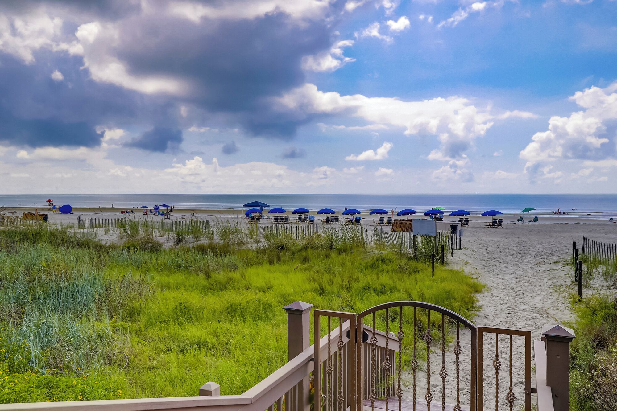 A beach with umbrellas and chairs, a wooden fence, and a grassy area in the foreground.