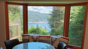 A dining area with a round glass table surrounded by six chairs, large windows offering a view of a forest and a lake, and a ceiling light fixture. - #Slowtravel+Wellness~woodfired hot tub, cold plunge, sauna, forestbathing, beach (Halfmoon Bay)