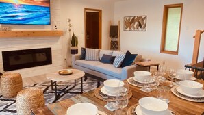 A modern living room with a fireplace, a sofa with patterned cushions, and a dining area set for a meal. - #Slowtravel+Wellness~woodfired hot tub, cold plunge, sauna, forestbathing, beach (Halfmoon Bay)