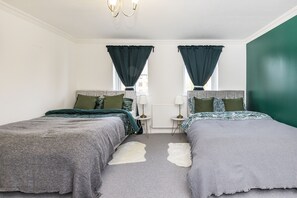 A bedroom with two beds, a small table, and a window with curtains. - Inglewood Hub London (London)