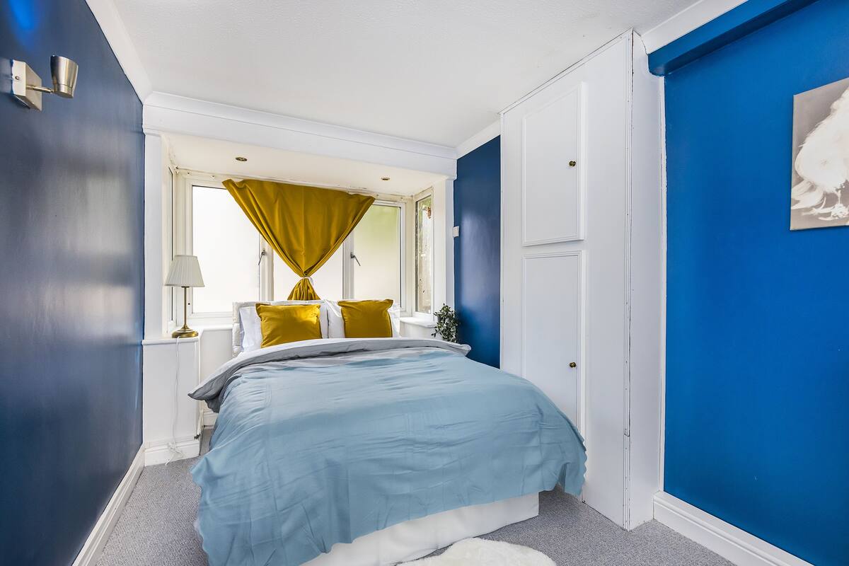 A bedroom with a bed, a window with yellow curtains, a white door, and a blue wall.