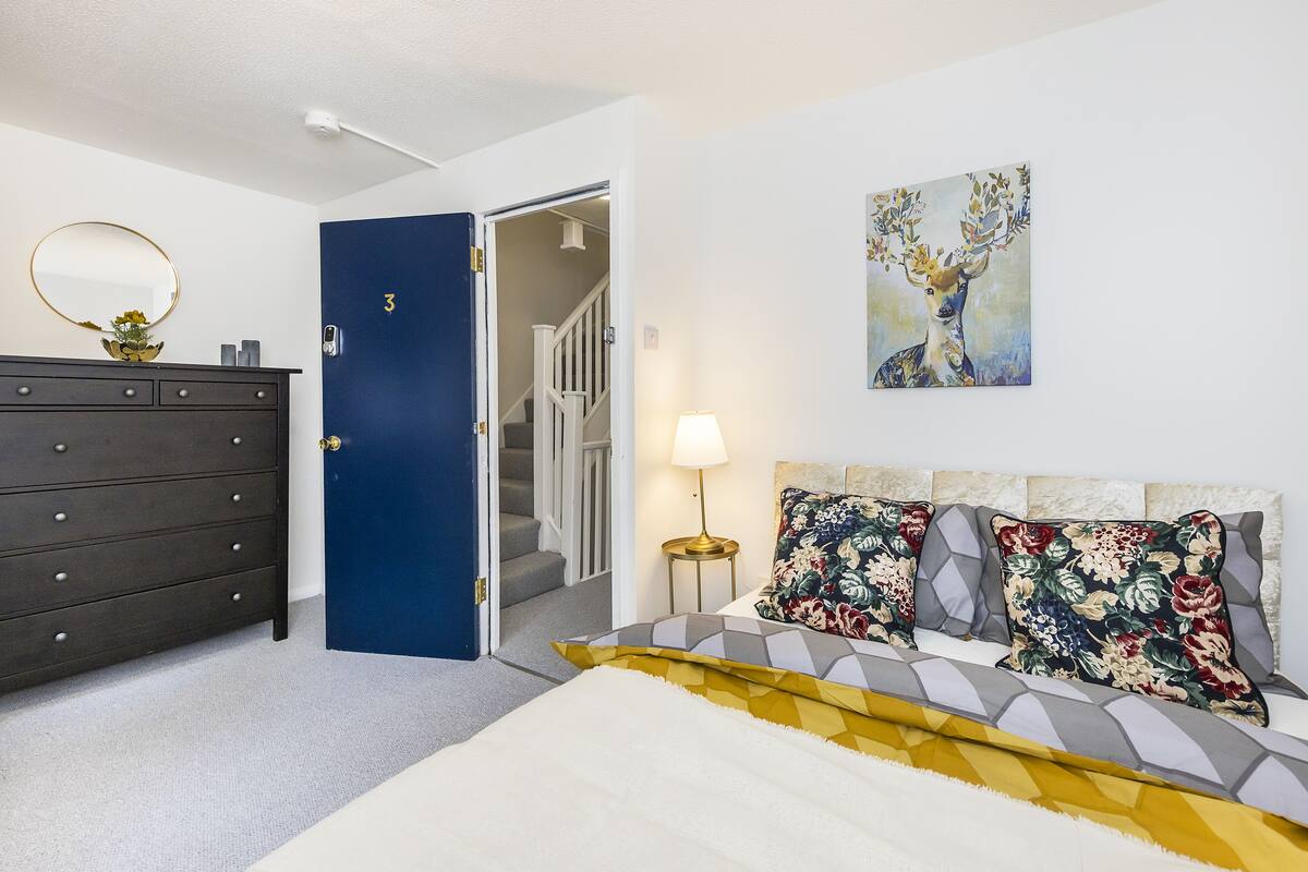 A bedroom with a bed, a dresser, a door, a staircase, and a painting on the wall.