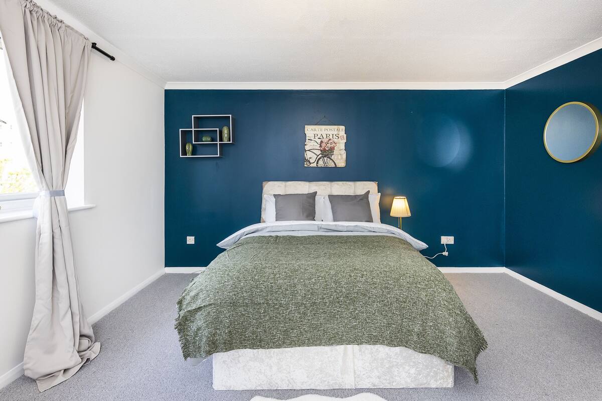 A bedroom with a bed, a nightstand, a lamp, a wall-mounted mirror, and a window with curtains.