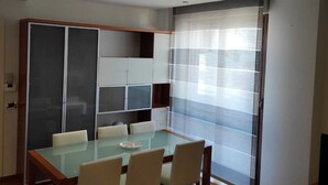 Ein moderner Essbereich mit Glastisch und Stühlen, einer integrierten Küchenzeile und einem großen Fenster mit transparenten Vorhängen.
