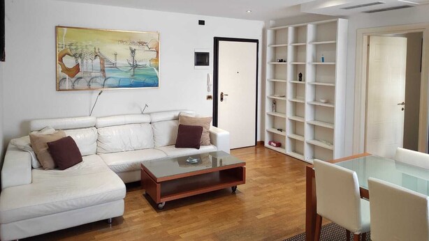 Ein modernes Wohnzimmer mit einem weißen Sofa, einem Glastisch und einem Essbereich mit Tisch und Stühlen. An der Wand hängt ein Gemälde, und eine Tür führt in einen weiteren Raum.