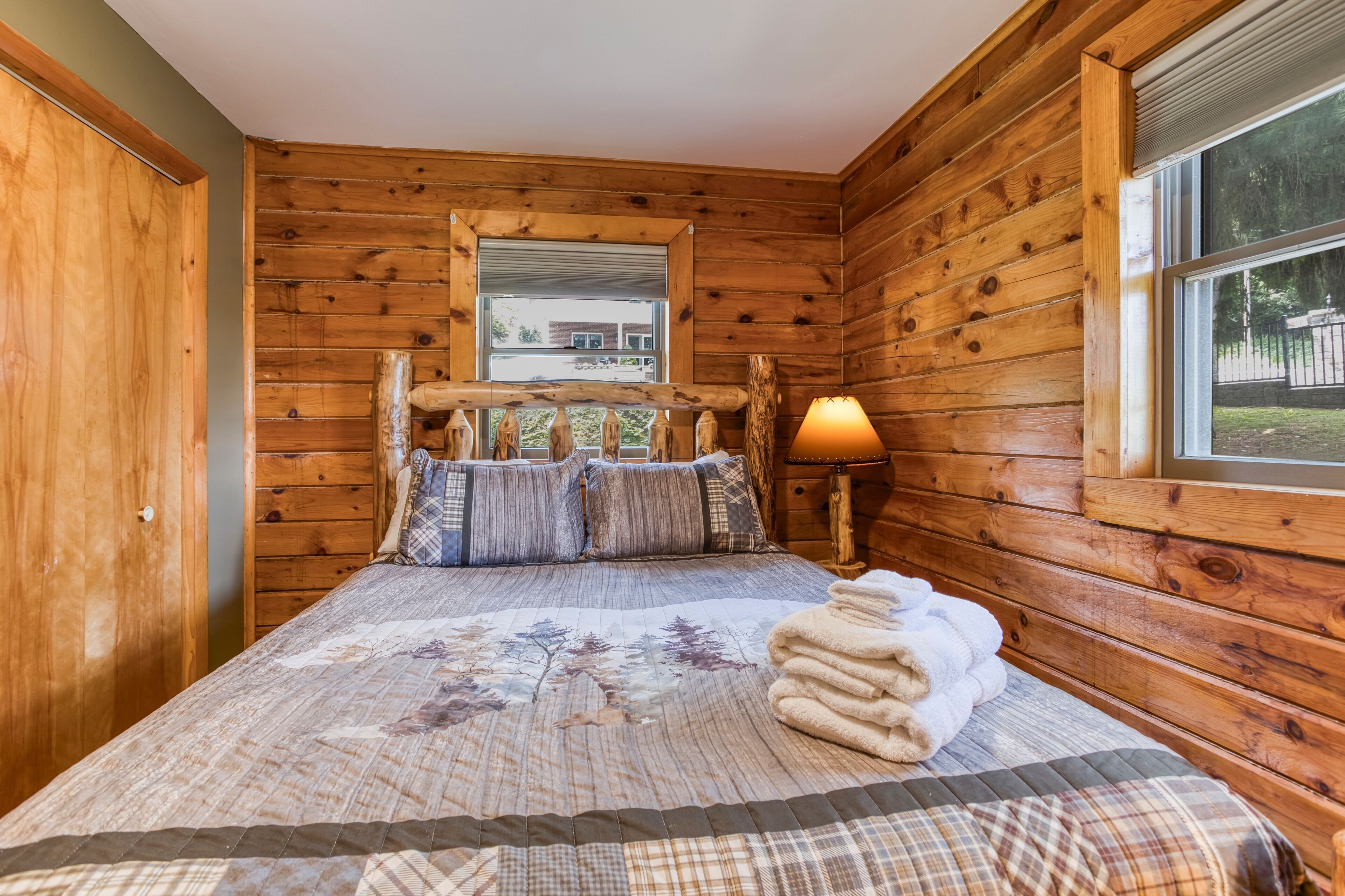 A wooden cabin bedroom with a bed, pillows, a lamp, and a window.