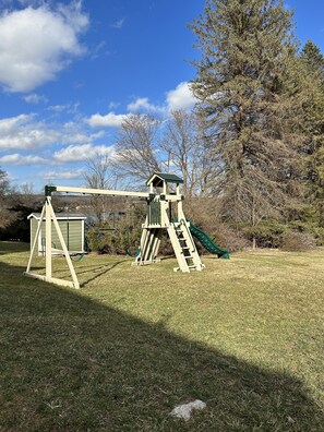 A wooden swing set with a slide, a small shed, and a tree in a grassy yard.