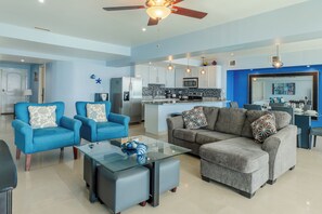 Ein modernes Wohnzimmer mit blauen Sofas, einem Glastisch und einem grauen Sofa mit gemusterten Kissen.