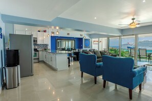 A modern kitchen with white cabinets, a stainless steel refrigerator, and a dining area with blue chairs and a view of the ocean.
