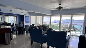 A modern living room with a large window offering a view of a cruise ship and ocean. There is a dining area with a bar and a ceiling fan.
