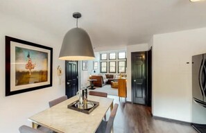 A modern kitchen with a dining area, featuring a large pendant light, a framed artwork, and a view into the living room.