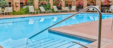 A swimming pool with a metal ladder, surrounded by lounge chairs and umbrellas, with a building in the background.