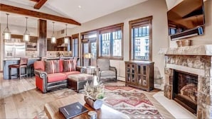A spacious living room with a fireplace, large windows, and a kitchen in the background. - Luxury 4 Bedroom Penthouse Rental off Main Street Within Walking Distance to Peak 9 (Breckenridge)